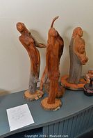 Four carved cedar statues on a blue surface with labels. The statues depict elongated female human forms with varying poses and detail. They are mounted on round wood bases with bark edges.