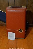 Brown hard carrying case standing upright, with a note 'Lot 25 Vintage Silver Projector'