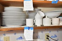 Shelf displaying stacked white dinner plates, smaller plates, several cups with handles, sugar bowl with lid, and a small bowl, all white ceramic with subtle embossed patterns.
