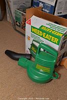 Green electric weed eater blower with black nozzle, boxed, sitting on carpet.