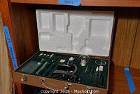 Open wooden box showing vintage chemistry set components including a black microscope and various small glass containers and tools fitted in green felt.