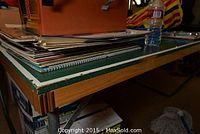 Photo of the corner of the green ping pong table showing wooden legs and edge wear, items stacked on top not included.