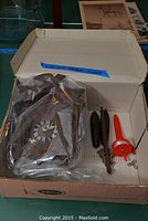 Wooden cuckoo clock wrapped in plastic inside cardboard box, with pine cone weights and winding key beside it.