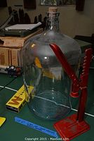 Large clear glass jug with cork and red metal wing-style wine opener on a table