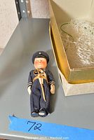 Small vintage soldier doll dressed in navy blue uniform with white gloves and a sword, displayed with a white and gold-trimmed box with packing material.