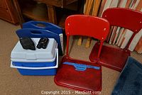 Photo showing two red metal stadium folding seats, one blue plastic stadium folding seat, blue and white cooler, and binoculars with carrying case on top of cooler.