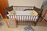 Wide shot showing the wooden baby crib with white lining and two cushions, the safety gate, and folded highchair on the floor.