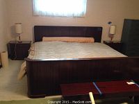 Front view of dark wood bed frame with mattress and bench; lamps and other furniture in background