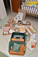 Photo showing the green case Phoenix sewing machine with wooden base surrounded by various sewing notions including threads, boxes of pins, lace, and sewing tools.