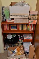Bookshelf with vintage books on shelves and multiple boxes and miscellaneous items on top, including some cardboard boxes and paper items.