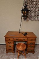 Full view of wood desk and small wicker stool. Vintage hanging lamp suspended above desk with chain. Desk has nine drawers with brass pulls.