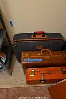 Three luggage pieces shown together against a wall; two vintage hard-shell suitcases and one fabric suitcase with orange and black color scheme.