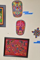 View of two Huichol yarn art masks and a framed yarn painting on wall with bright detailed colors and tribal patterns.