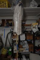 Side view photo showing the patio umbrella closed and covered in plastic, mounted on a beige pole with crank handle, standing in a square base.