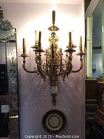 Ornate brass-tone metal electric wall candelabra mounted on wall; seven arms with bulb sockets
