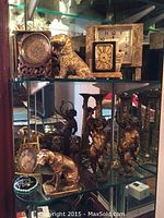 Glass display shelving unit with three glass shelves holding various decorative items including decorative plate on wood stand, brass dog figurine, and large marble clock