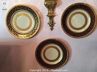Three gilt floral motif porcelain plates with black border displayed on wall with gold decorative wall sconce between. Plates are 10 3/4 inches in diameter and have gold intricate floral patterns around the rim.