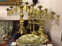 Four brass menorahs and one tall brass candle holder arranged on a table alongside glassware