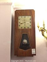 Full view of the wooden Art Deco wall clock showing the dial, pendulum window, and stepped geometric design.