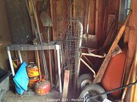 Full view of the corner shelf holding chicken wire rolls, vintage gas cans, and garden tools leaning against the wall.