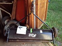 Full view of the Blue Hawk lawn sweeper attachment showing black metal frame, wheels, and manual instructions taped on frame.