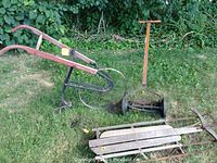 Manual cultivator featuring red wooden handles and black metal frame with a large spoked wheel, set in grass near a bushy area.