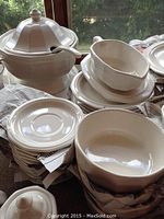Stacked white ceramic dinnerware including plates, bowls, gravy boat and a soup terrine with lid on a table near window.