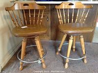 Two wood framed swivel bar stools with spindle backrests, metal foot bars, and vinyl seats with visible tears, shown side by side.