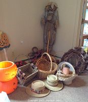 Photo showing collection of various baskets in different shapes and sizes, two straw hats, a large scarecrow figure standing against the wall, and stacked orange pumpkin-shaped plastic containers.