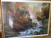Framed puzzle showing stone cottage with waterwheel and stream in a wooded setting, warm evening lighting