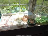 Full view of assorted glassware items on window ledge including pink depression glass, white and milk glass, green FireKing pieces.