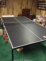 Full view of the black QuickPlay ping pong table with net, resting on a green carpeted floor in a wood-paneled room, showing the table is large, sturdy with metal legs.