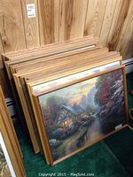 Collection of 12 framed Thomas Kinkade puzzles stacked together, showing frames and one visible scenic puzzle.
