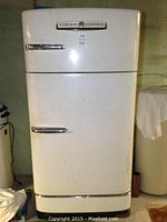 Full view of off-white vintage General Electric refrigerator showing two doors with chrome handles, standing in basement surroundings.