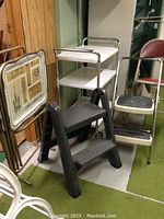 Wide view showing metal cart with three shelves, black step stool in front, metal TV trays on left, and metal folding chair on right