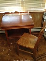 Photo showing wooden drafting desk with slanted hinged top, closed, and matching wooden chair beside it. Both show wear and age.