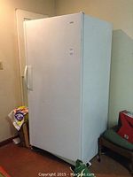 White upright freezer standing in a room next to a chair and some boxes. Appears clean with a slight textured finish.