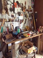 Wide view of a wooden work table covered with assorted tools including splitting mauls, portable lights, a vintage hand plane, with pegboard behind holding various hand tools.