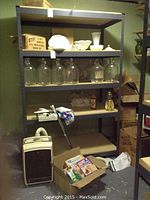 Front view of one shelving unit showing five shelves with various items present, including glass bottles and decorative glassware; metal frame is blue-gray.
