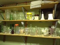 Shelf of assorted vintage glassware including clear and colored vases, pitchers, a small green glass plant frog, and various glass containers