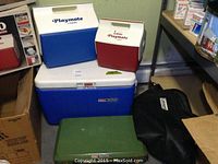 Two small Igloo Playmate coolers (blue/white and red/white), a large blue Coleman cooler, green Coleman propane camp stove, and black carrying case