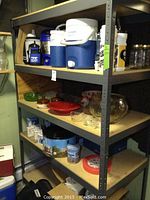 Metal shelving unit holding multiple Coleman coolers, assorted travel cups, glass serving bowls, punch bowl set, tins, and other kitchen items.