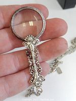 Close-up of the magnifying glass pendant showing detailed ornate filigree metalwork in the Art Nouveau style held in hand.