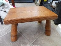 Side angle of the oak footstool showing the rectangular top and one turned leg, sitting on tiled floor.