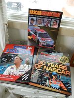 Three large hardcover NASCAR themed coffee table books titled NASCAR Superstars, NASCAR: A Celebration, and 50 Years of NASCAR, displayed on a table near a window.