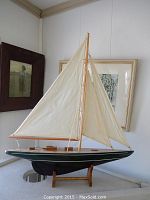 Full view of model pond sailboat on wooden stand showing cream fabric sails and dark green hull.