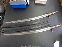 Set of two Asian military dress type swords with their leather scabbards laid side by side showing full length and details of blades and handles.