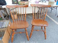 Set of two mid century wooden chairs made of maple with spindle backs and contoured seats, shown standing on pavement with other items nearby.