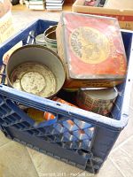 Blue plastic crate filled with vintage advertising tins and bottles including round and rectangular tins with colorful labels.