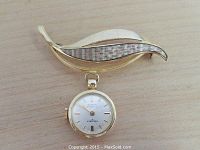 Gold brooch with a leaf-shaped design and a small round watch pendant attached, placed on a wooden surface.